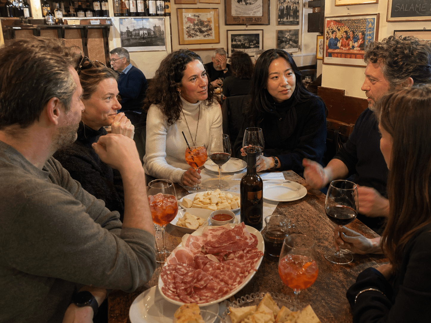 Amici seduti a un tavolo di legno gustano vino, spritz e taglieri in un'enoteca rustica.