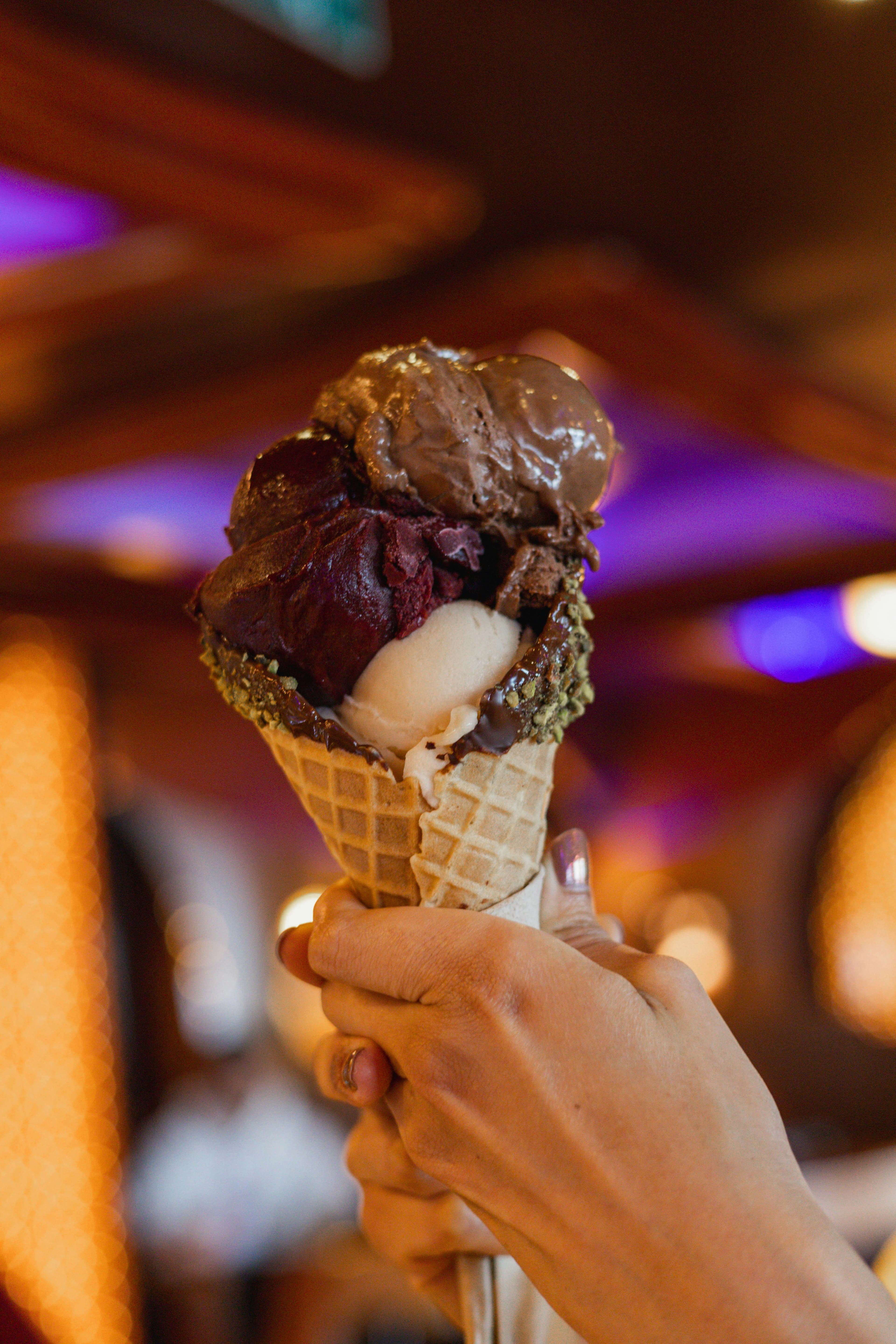 Mano che tiene un cono gelato con tre gusti e bordo al cioccolato e pistacchio.