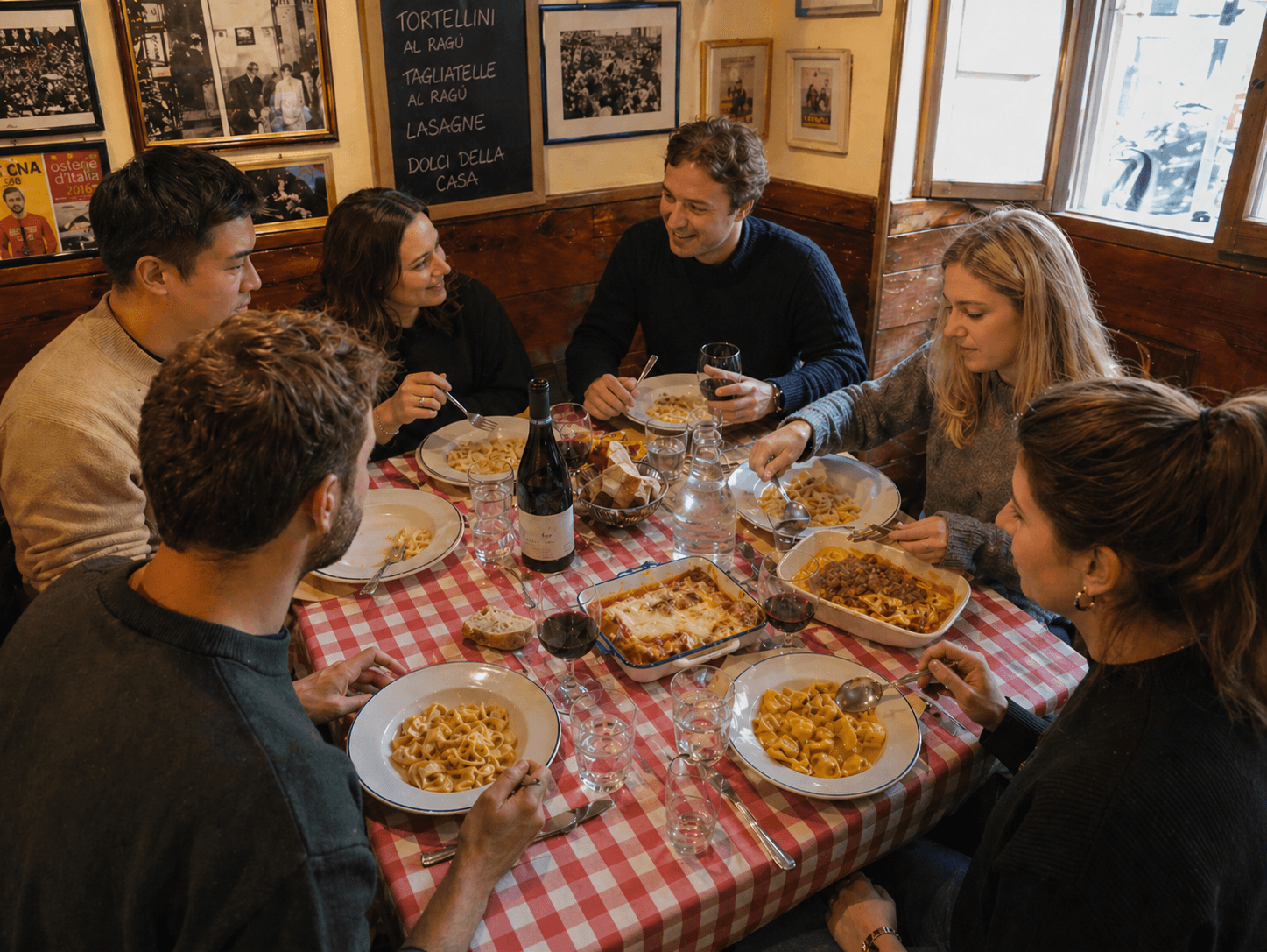Amici seduti a tavola mangiano pasta in una trattoria con tovaglia a scacchi rossi.